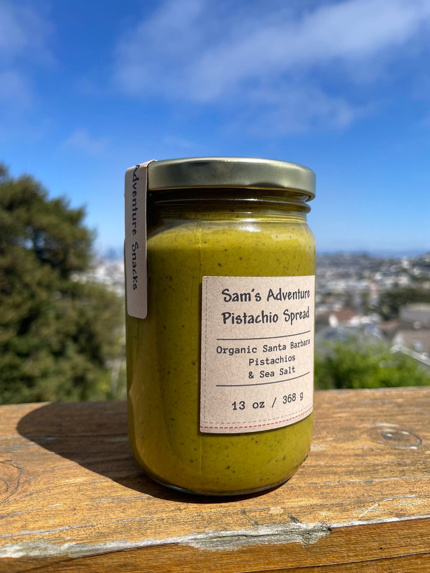 13 oz jar of organic santa barbara pistachio spread. Jar is placed on a wood bench with the city of San Francisco in the background. Label on jar lists "Sam's Adventure Pistachio Spread", "Organic Santa Barbara Pistachios & Sea Salt" and 13 oz/368g