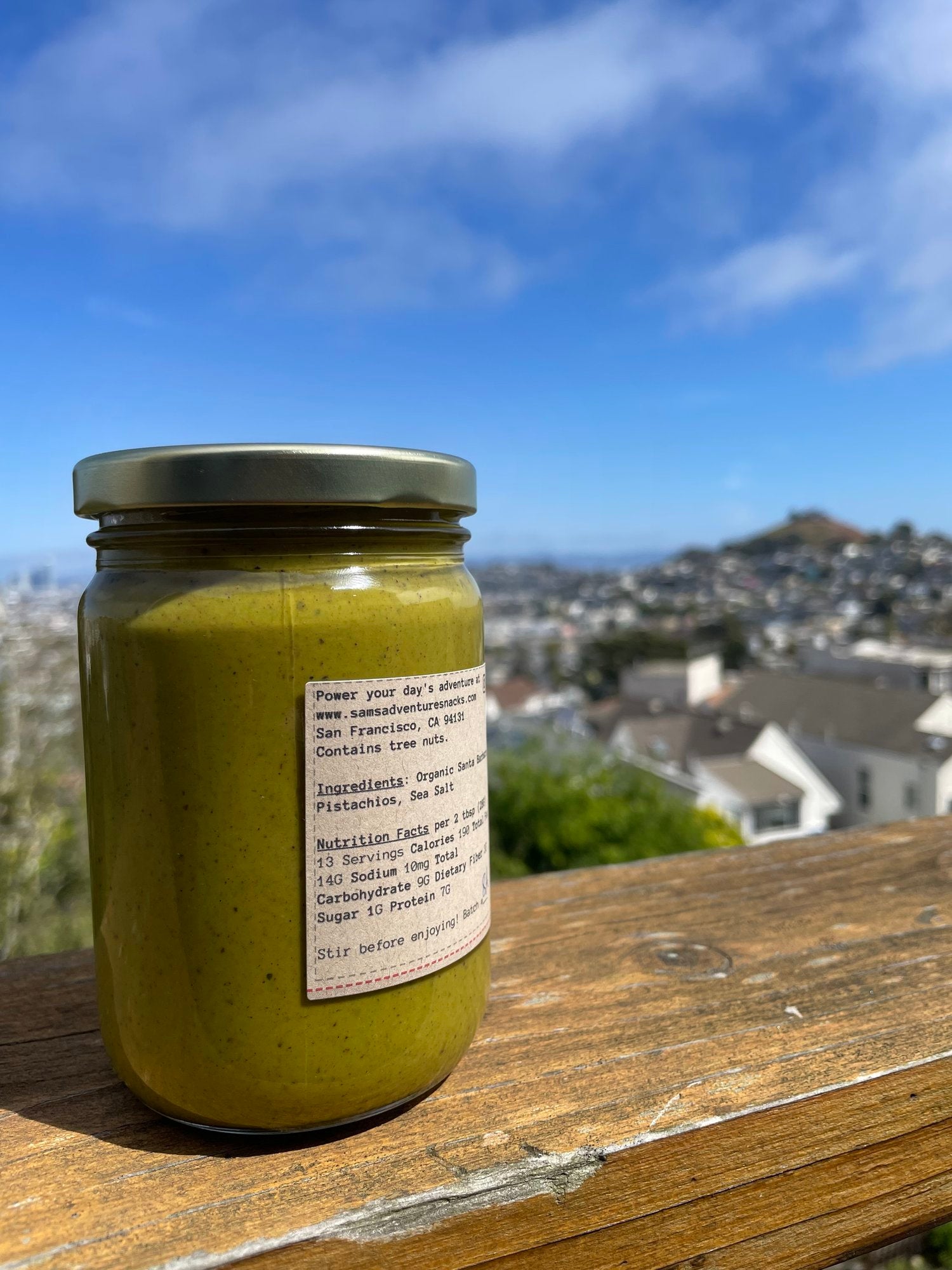 13 oz jar of organic santa barbara pistachio spread. Jar is placed on a wood bench with the city of San Francisco in the background. Jar is turned around to show back label with product description and ingredients.